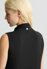 Zwarte mouwloze top met een hoge kraag, voorzien van een klein circulair metallic detail aan de achterkant, gladde textuur en aansluitend ontwerp.