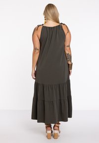 Dunkelgrünes Maxikleid mit verstellbaren Schulterbändern und gestuften Rüsche-Design, aus weichem Baumwollstoff gefertigt. Tragen mit Absatzsandalen.