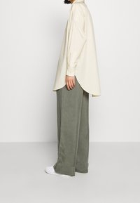 Chemise oversize crème à manches longues, ourlet arrondi ; associée à un pantalon ample vert olive et des baskets blanches. Tissu lisse, coupe décontractée.