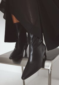 Botas de cuero negro de punta afilada y tobillo con un diseño ajustado y un tacón esbelto, exhibidas sobre un taburete metálico contra un fondo neutro.