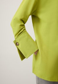 Roheline puuvillane särk, millel on lai ülespööratud mansett kuldse nupuga, sile tekstuur ja talje juures nähtav küljetasku.