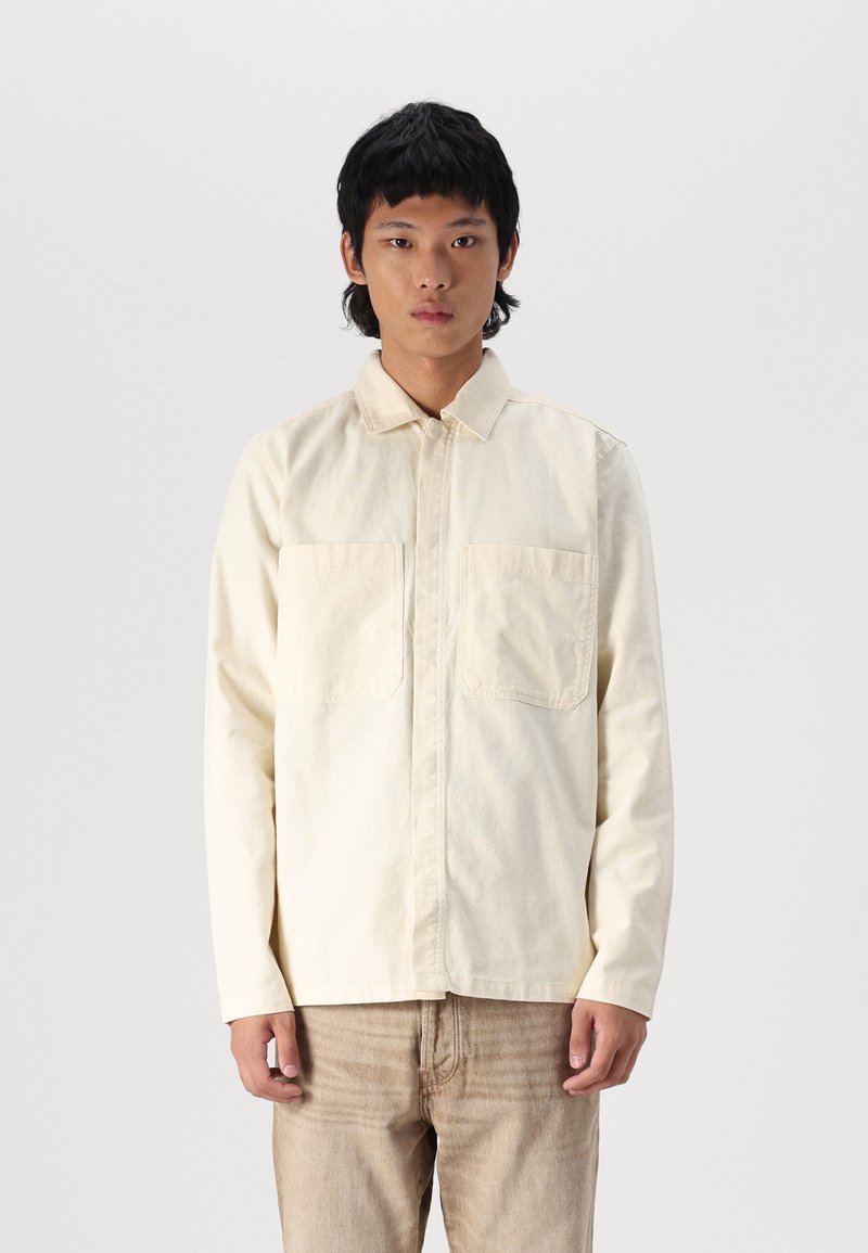 Light beige long-sleeve shirt with a collared neck, two chest pockets, and a straight cut, made from a textured cotton fabric.