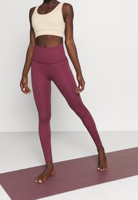 Leggings bordeaux a vita alta abbinati a un crop top beige. I leggings presentano una texture liscia e dettagli di cucitura sottili.