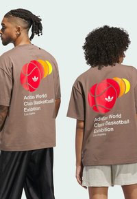 Hnedé bavlnené tričko s viacfarebnou grafikou basketbalového lopty na chrbte a textom "Adidas World Class Basketball Exhibition" pod ním.