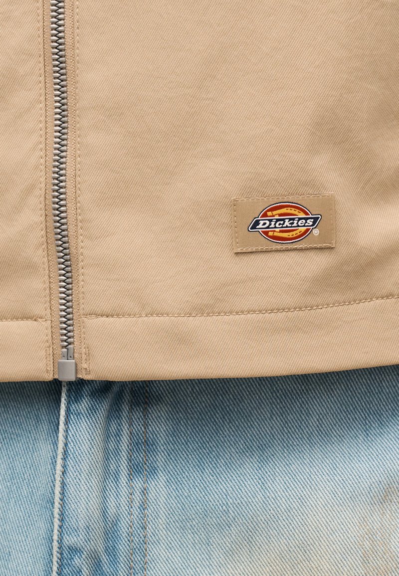 Beige Jacke mit silbernem Reißverschluss, teilweise geschlossen, die das Dickies Logo-Patch zeigt, getragen über hellblauen Jeans.