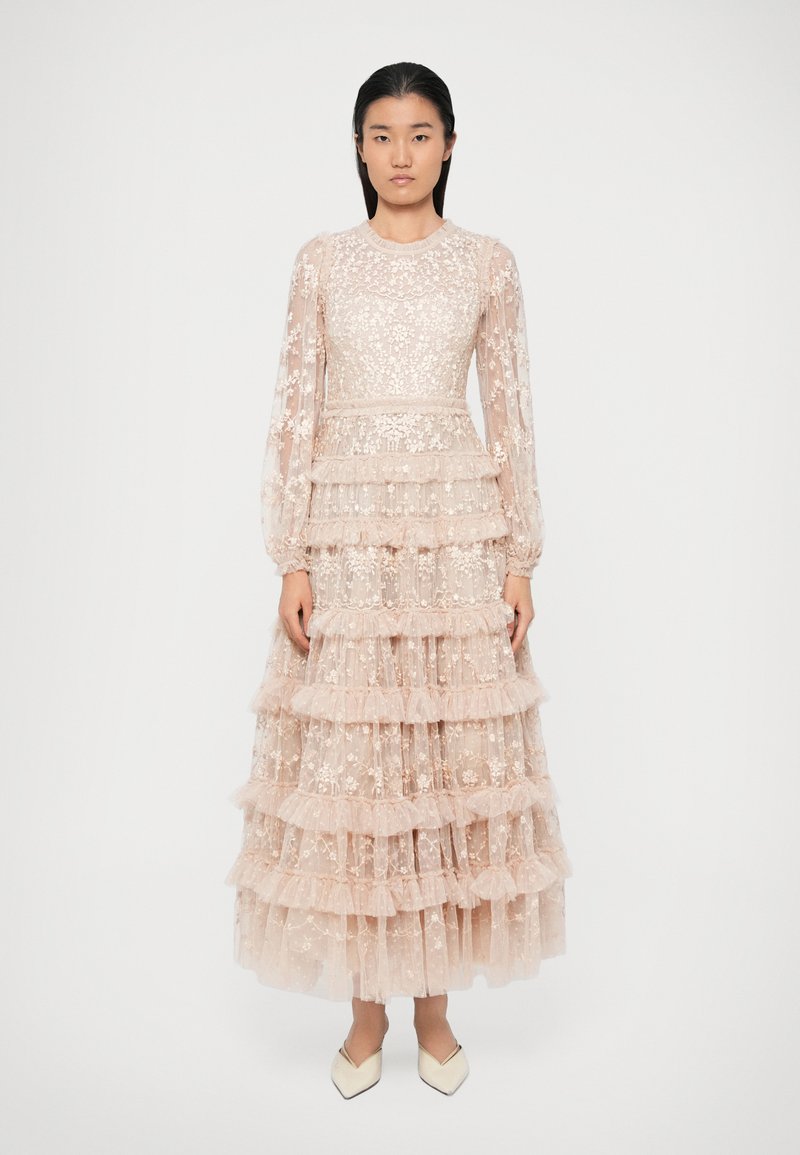 Švelniai rožinė nėriniuota suknelė su ilgomis rankovėmis, pakopinėmis volanais, gėlėmis siuvinėta ir prigludusia liemene. Ruošiniai kreminiai bateliai puikiai papildo aprangą.