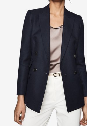 Kobieta ubrana w granatową, dwurzędową marynarkę na satynową taupeową bluzkę, zestawioną z białymi spodniami i jasnym paskiem.