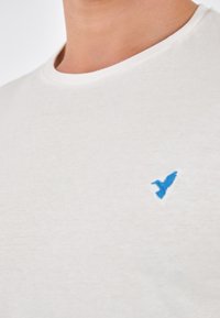 T-shirt in cotone beige chiaro con colletto a girocollo e un piccolo logo di un uccello ricamato in blu sulla parte sinistra del petto.