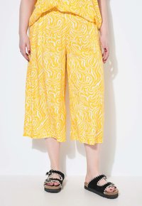 Gelbe Culotte-Hosen mit weißen abstrakten Mustern, kombiniert mit schwarzen Rutschschuhen mit dekorativen Akzenten und einer strukturierten Sohle.