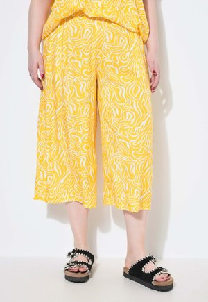 Pantalones culotte amarillos con patrones abstractos en blanco, combinados con sandalias deslizables negras que presentan acentos decorativos y una suela texturizada.