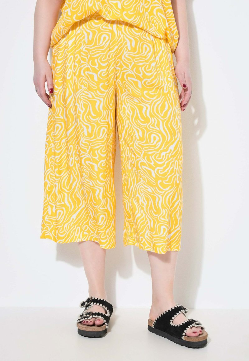 Gelbe Culotte-Hosen mit weißen abstrakten Mustern, kombiniert mit schwarzen Rutschschuhen mit dekorativen Akzenten und einer strukturierten Sohle.