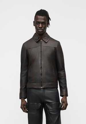 Μαύρος άνδρας φοράει σκούρο καφέ δερμάτινο μπουφάν με φερμουάρ και μαύρο δερμάτινο παντελόνι με μεγάλες μπροστινές τσέπες, στέκεται μπροστά σε απλό φόντο.