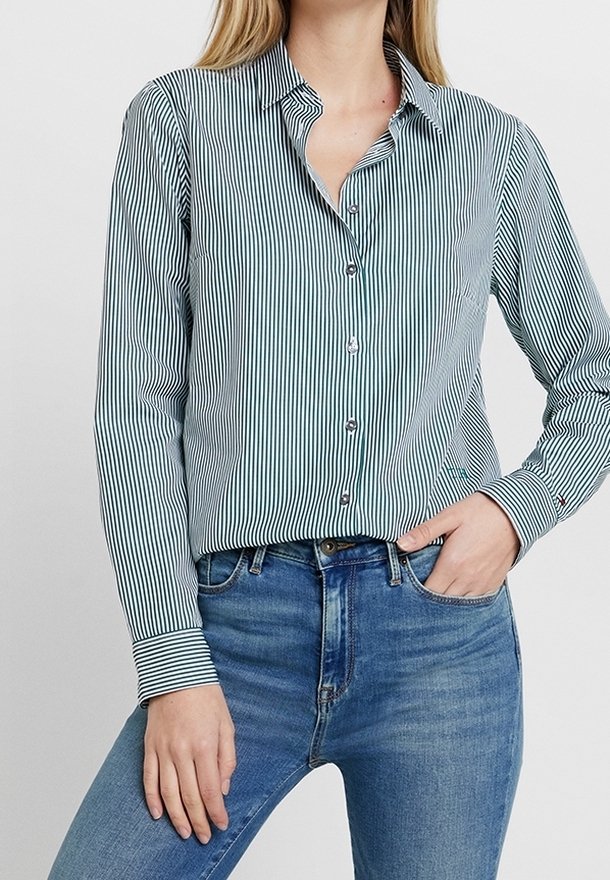 Femme portant une chemise à rayures verticales bleu et blanc boutonnée, rentrée dans un jean bleu moyen, une main dans la poche.