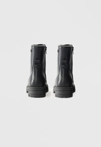 Botas de tornozelo em couro preto com uma textura lisa, solas de borracha grossa, zíperes laterais e detalhes de tachas ao longo das bordas superiores.