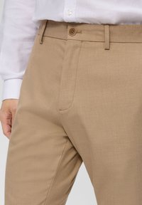 Beige trousers with a smooth texture, featuring a front pocket, belt loops, and a single button closure. Paired with a white long-sleeve shirt.