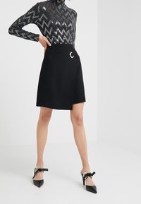 Svart invecklad kjol av ett mjukt tyg, med en silverfärgad halvmånsdetalj. Modellen bär en glittrig svart zigzag-topp och spetsiga klackar.