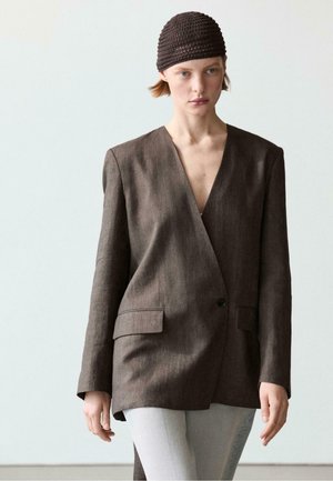 Kobieta w teksturowanej brązowej oversize marynarce, jasnoszarych spodniach i ciemnobrązowej dzianinowej czapce, stojąca na tle jednolitego jasnego tła.