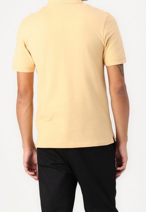 Jasnożółta koszulka polo z krótkim rękawem, wykonana z gładkiej tkaniny. Posiada boczne rozcięcia u dołu, noszona z czarnymi spodniami.