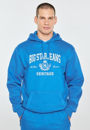 Mężczyzna o krótkich włosach, ubrany w niebieską bluzę z kapturem z napisem "Big Star Jeans Heritage" oraz pasujące niebieskie spodnie, ręce w kieszeniach, neutralne tło.