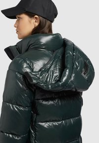 Donkergroene, glanzende pufferjack met een hoge kraag en een afneembare capuchon. Voorzien van een rits en stofaccenten, met een stijlvolle, doorgestikte design.