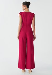 Červený overal s širokými nohavicemi, přiléhavým pasem a krátkými rukávy. Hladká látka se zadním zipem a dekorativním švem.