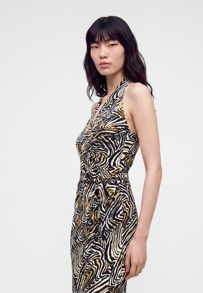 Femme aux longs cheveux noirs portant une robe sans manches à motifs avec des rayures abstraites noires, blanches et dorées, similaires à celles d’un zèbre.
