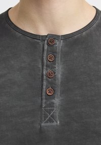 Grau Baumwoll-Henley-Shirt mit Rundkragen, vier braunen Knöpfen und kontrastierenden Nähten. Glatte Textur mit einem verblassten Finish.