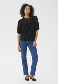 Sort bluse med broderede detaljer, puffærmer og en struktureret pasform, parret med blå straight-leg jeans og sorte hæle.