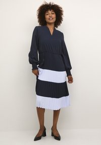 Jūras spēku un baltas krāsas bloku kleita ar plīsumiem, garām pūkainām piedurknēm un V veida kakla izgriezumu. Sānu šķēlumi kustību ērtībai.