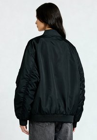 Zwarte bomberjas met een gladde, glanzende textuur, geribbelde manchetten en tailleband. Heeft geplooide zijnaden en een opstaande kraag.