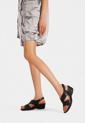 Donna che indossa pantaloncini cargo argento lucidi e sandali con zeppa neri, mostrando gambe e parte della mano su sfondo bianco.