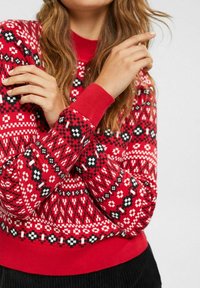 Pull rouge en tricot avec un motif géométrique en blanc et noir, doté de poignets et d'un ourlet côtelés, affichant une finition texturée.