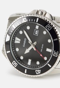 Montre Casio avec un cadran noir, un boîtier en acier inoxydable et des marqueurs lumineux. Elle présente une lunette argentée, un guichet de date et une aiguille des secondes rouge.