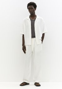 Camicia bianca a trama e pantaloni larghi abbinati con elastico in vita, sovrapposti a una maglietta grigia, abbinati a ciabatte nere e accessori in argento.