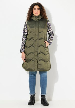 Olivengrüne gepolsterte Weste mit hohem Kragen und Zickzackmuster, ausgestattet mit seitlichen Taschen. Getragen über einem blumigen Langarmoberteil und blauen Jeans.