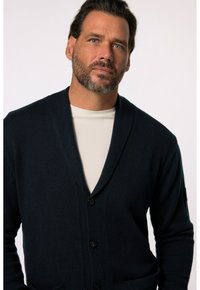 Marinblå stickad kofta med sjalkrage, prydd med fem svarta knappar och sidofickor. Under finns en ljusfärgad skjorta synlig.