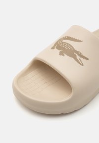Sandală din cauciuc bej, cu o talpă texturată, design cu vârf deschis și un logo de crocodil auriu, contrastant, imprimat pe partea superioară.