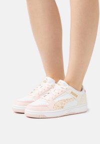 Baskets roses et blanches avec un accent texturé beige, ornées de marquages dorés et d'une fermeture à lacets. Le design comprend des détails perforés sur l'avant.