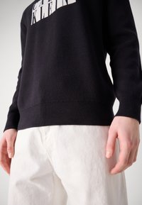 Black sweater with white lettering on the chest, knitted texture, ribbed cuffs and hem, paired with white pants. Close-up on hands.