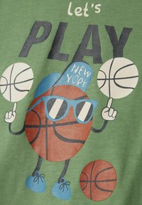 Una tela verde presenta un personaje de dibujos animados que se asemeja a una pelota de baloncesto, usando gafas de sol y una gorra azul con la inscripción "New York", y sosteniendo balones de baloncesto.