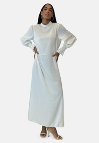 Robe longue blanche en tissu soyeux, avec des manches bouffantes ornées de smocks aux poignets et un col montant. Silhouette lisse et fluide.