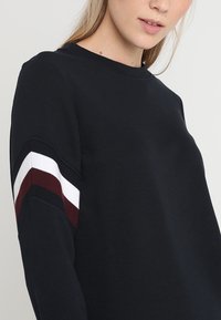 Mujer vistiendo una blusa de manga larga negra con rayas en burdeos, blanco y negro en la parte superior del brazo, cortada en el cuello y la torso.