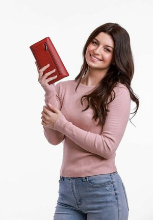 Červená kožená peněženka s texturovaným povrchem, zipovou kapsou a obdélníkovým tvarem, držena osobou v růžovém žebrovaném svetru.