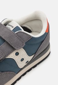 Grå og blå sneakers med overdel i semsket og tekstil, hvite logo aksenter, borrelåsrem, og oransjefarget hældetalj, med sort gummisåle.