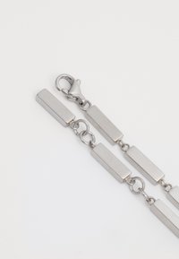 Bracciale in metallo argentato con anelli rettangolari e chiusura a granchio. La superficie è liscia e opaca con una tonalità di grigio uniforme.