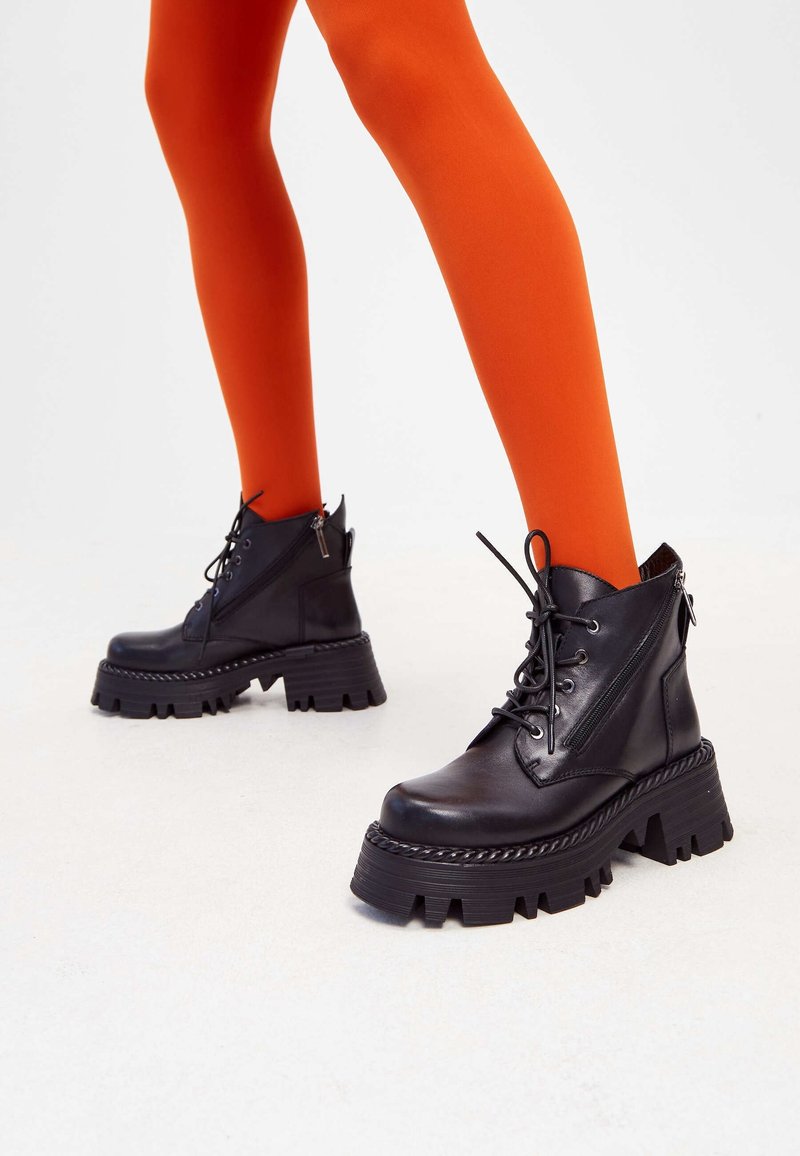 Jambes portant des collants orange vif associés à des bottes montantes à lacets en cuir noir épais avec des semelles épaisses et rainurées sur un sol blanc.
