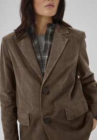 Blazer din suede maro cu un design elegant, două butoane și buzunare frontale, purtat peste o cămașă în carouri albastre și gri.