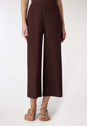 Donna indossa pantaloni marroni cropped a vita alta, a gamba larga e con texture, e sandali piatti dorati con cinturini alla caviglia, in piedi su sfondo bianco.