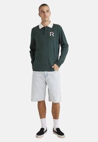 Dark green long-sleeve polo shirt with a white collar and embroidered logo, paired with light denim shorts and black slip-on shoes.