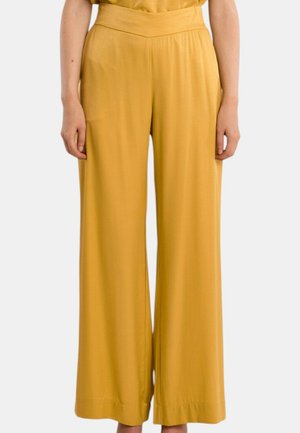 Gule højtaljede bukser med vidde ben, båret af en person, der står med armene afslappet ned langs siderne mod en ensfarvet baggrund.